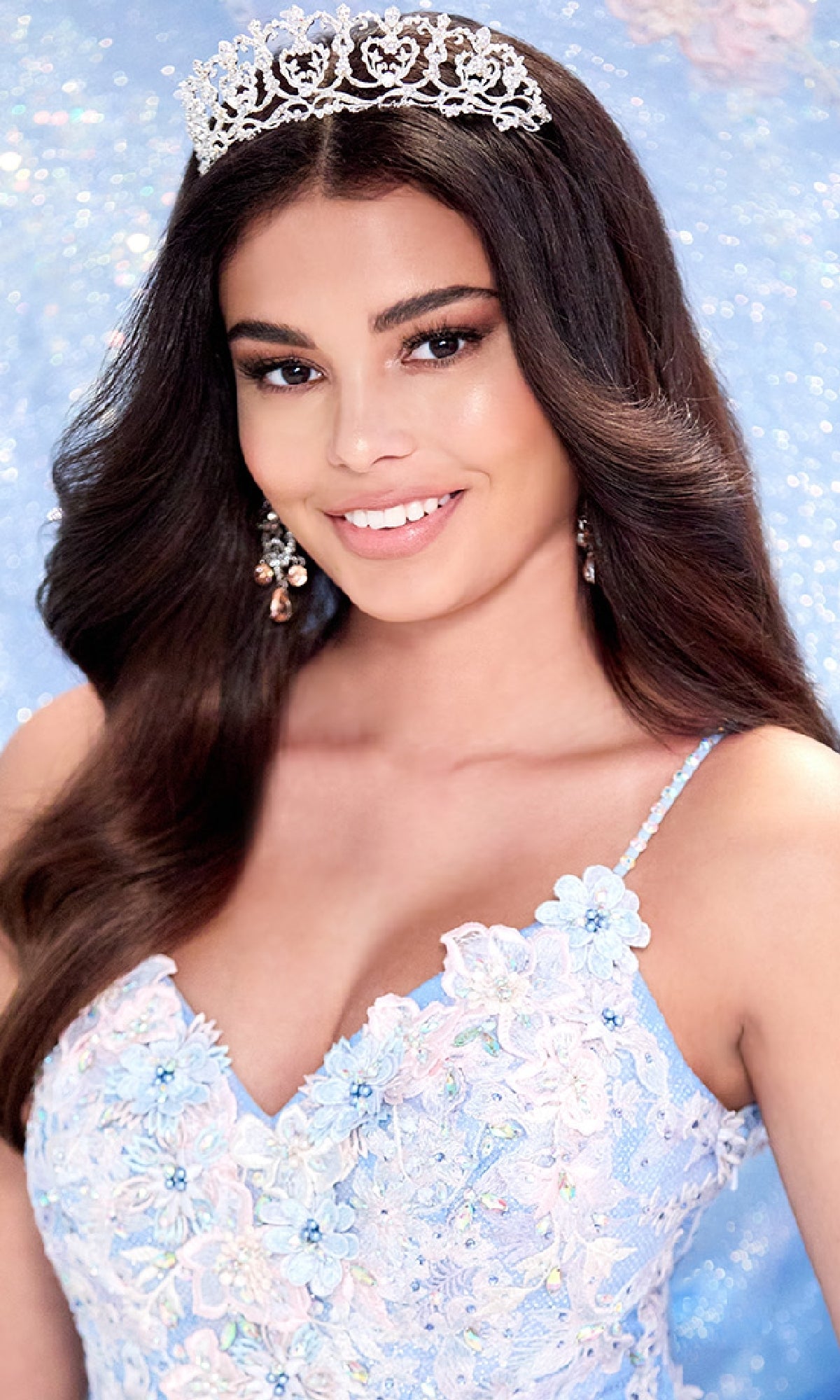 A woman with long brown hair wearing a silver tiara, drop earrings, and the Light Blue Quinceanera Dress: Princesa PR30083 with a corset bodice smiles against a sparkly blue background.